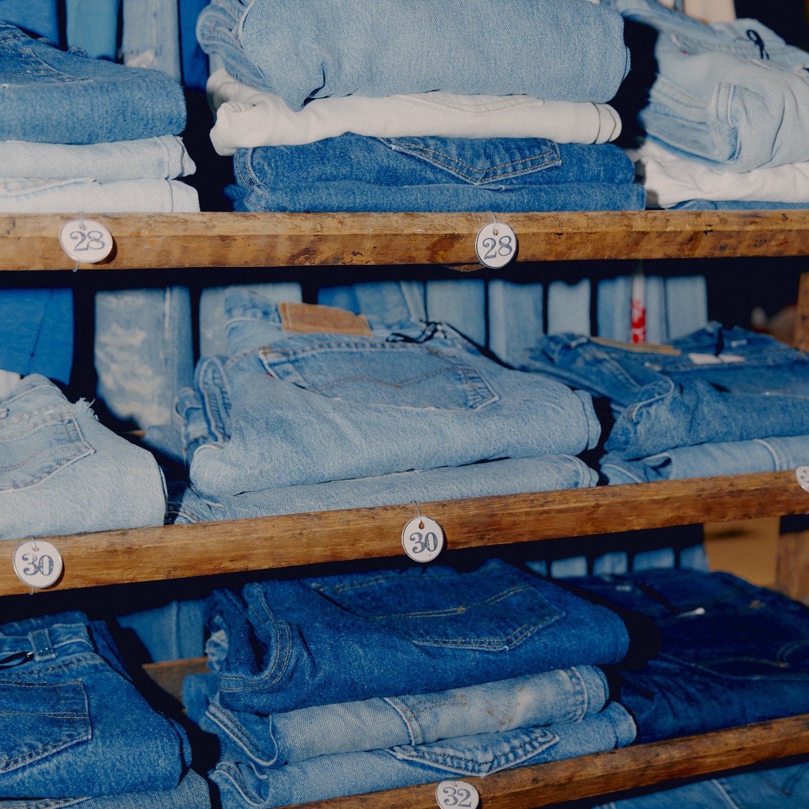 Stacks of folded blue denim jeans arranged on wooden shelves, with small white price tags attached to the shelves.