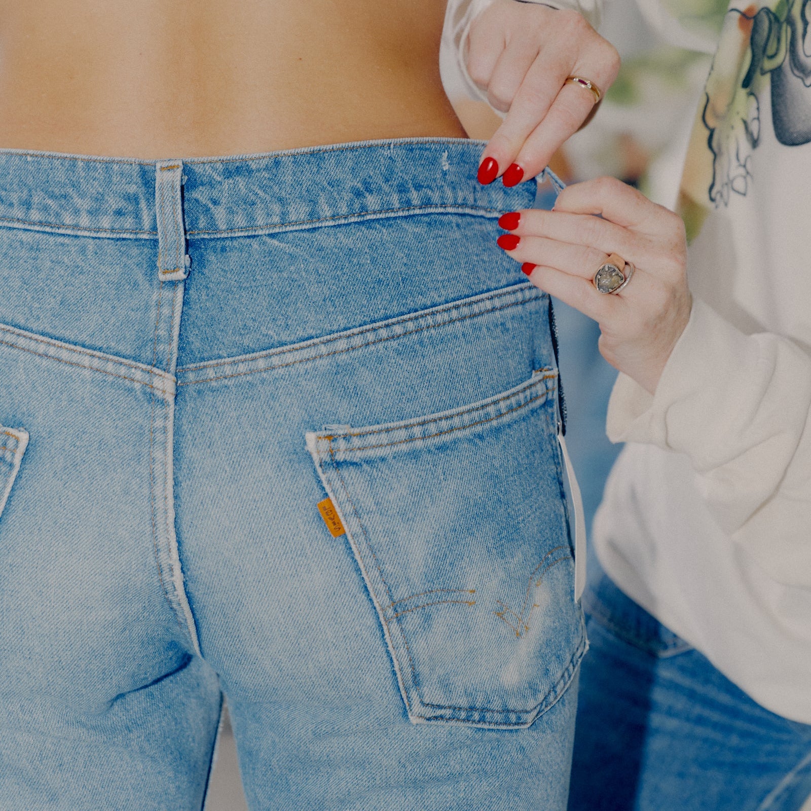Close-up back view of a person wearing light blue denim jeans, with another person adjusting the waistband. The photo focuses on the jeans, hands, nails, and part of a white patterned top.
