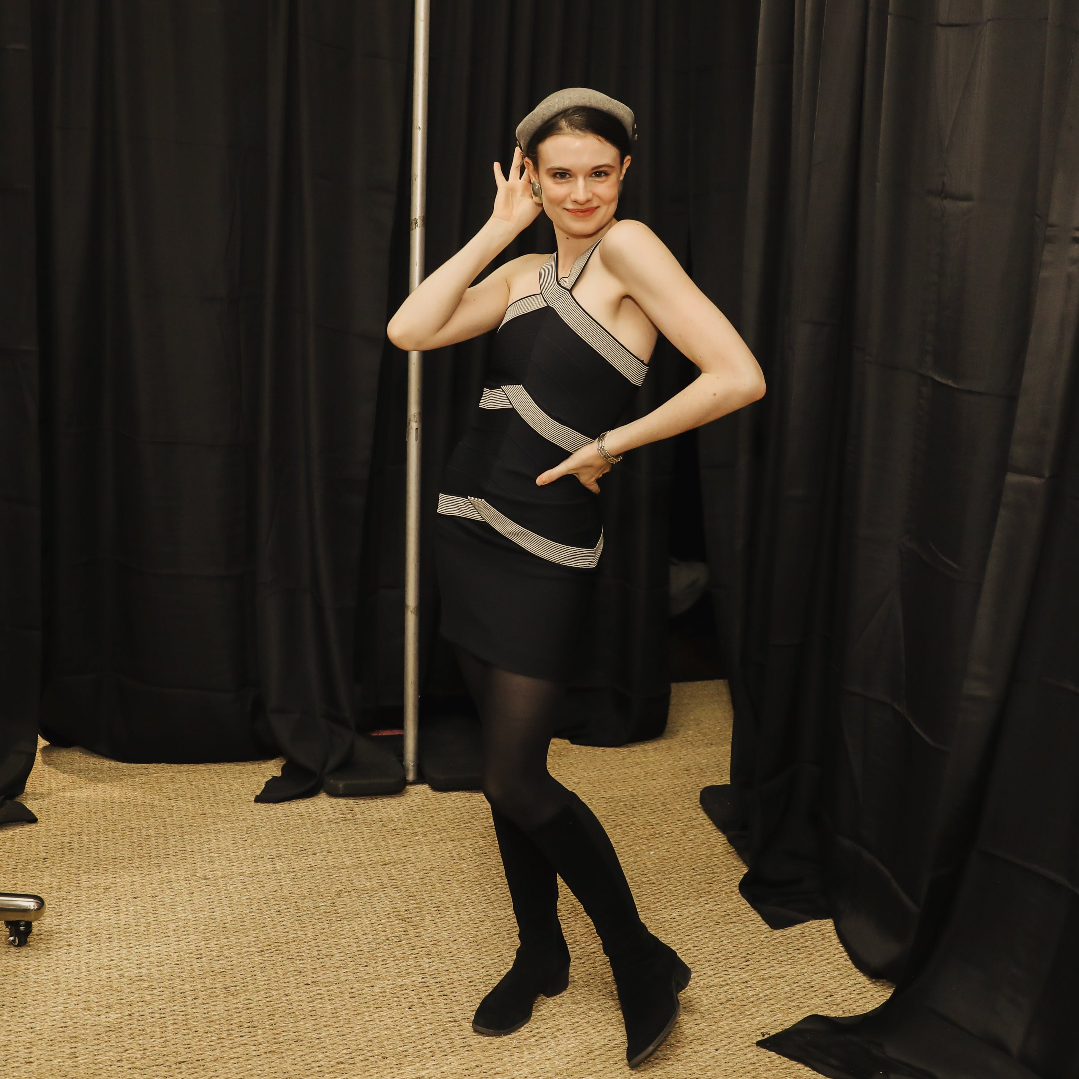 A woman modeling a black one-shoulder mini dress with silver-gray geometric stripes, paired with black tights and black shoes, posing in front of black curtains with a full-length mirror visible on the left.