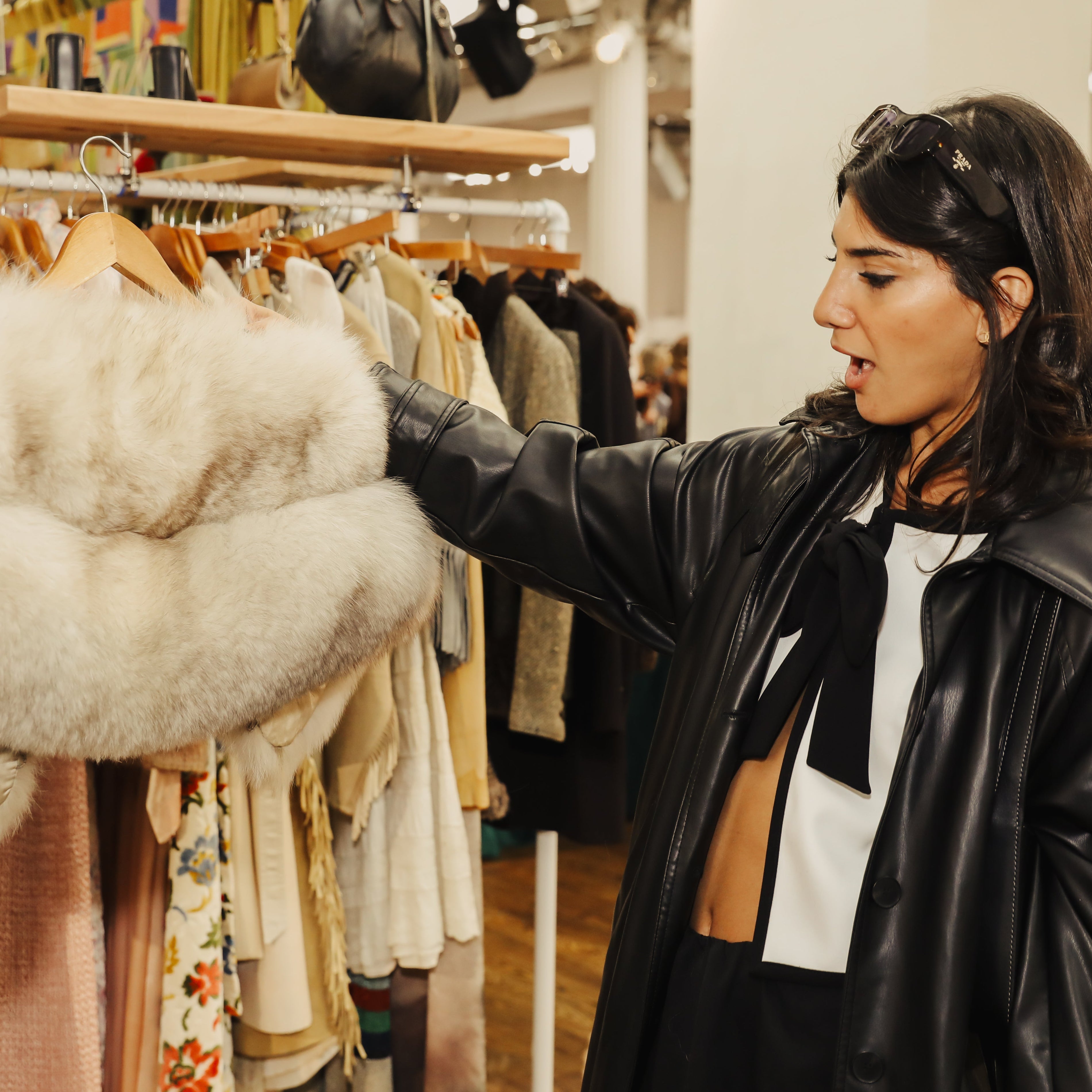 A woman in a black leather jacket browses a clothing rack in a boutique, holding up a fluffy cream faux fur collar.