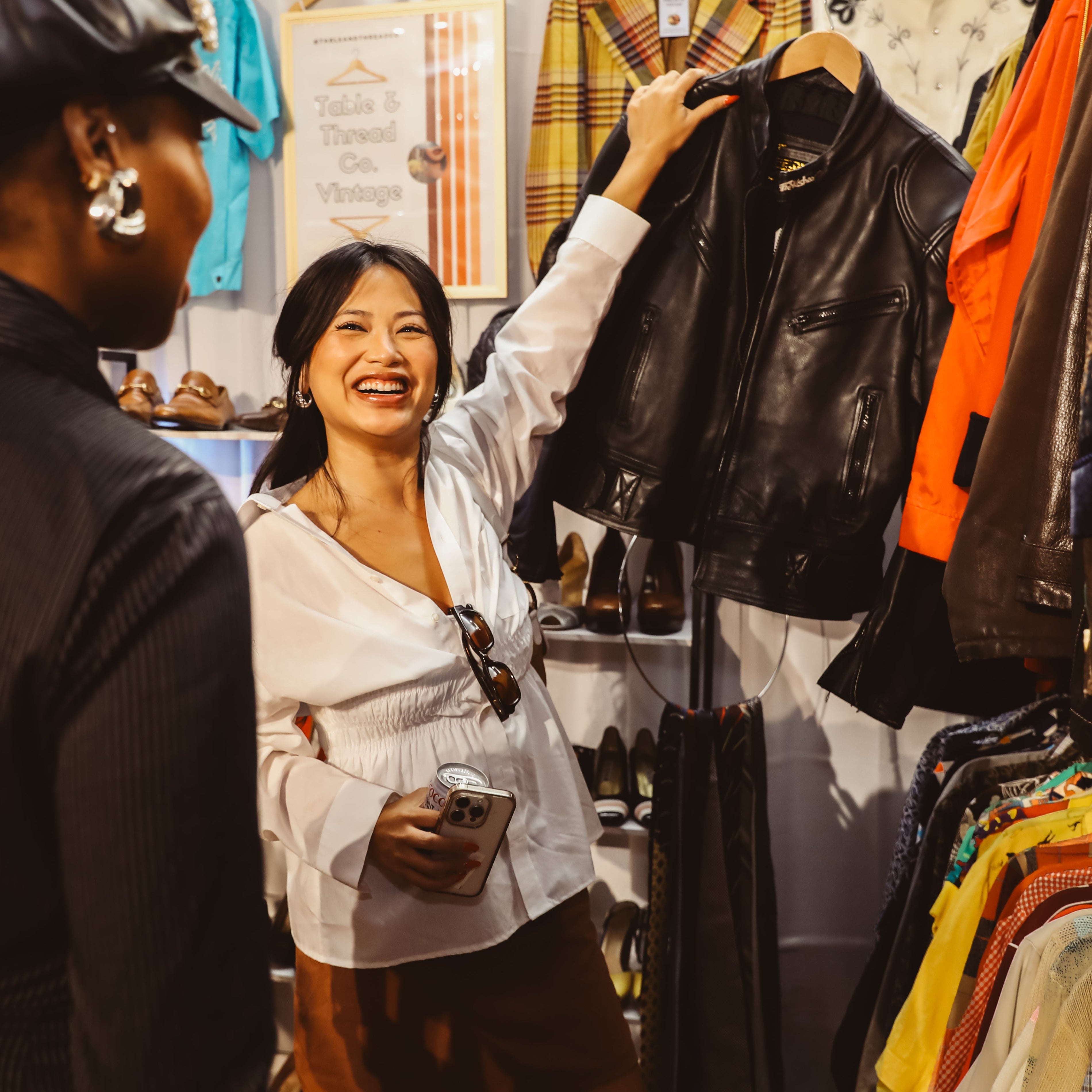 A woman in a white blouse smiles while holding up a black leather jacket on a hanger inside a clothing boutique, with another person in the foreground and racks of garments visible.
