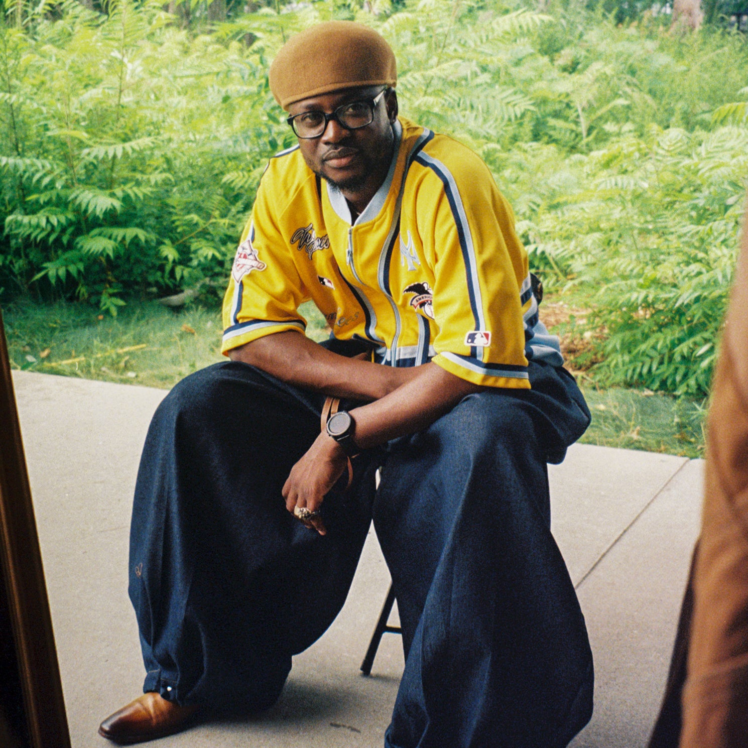 Person wearing a yellow jacket and blue pants sitting outdoors with greenery in the background