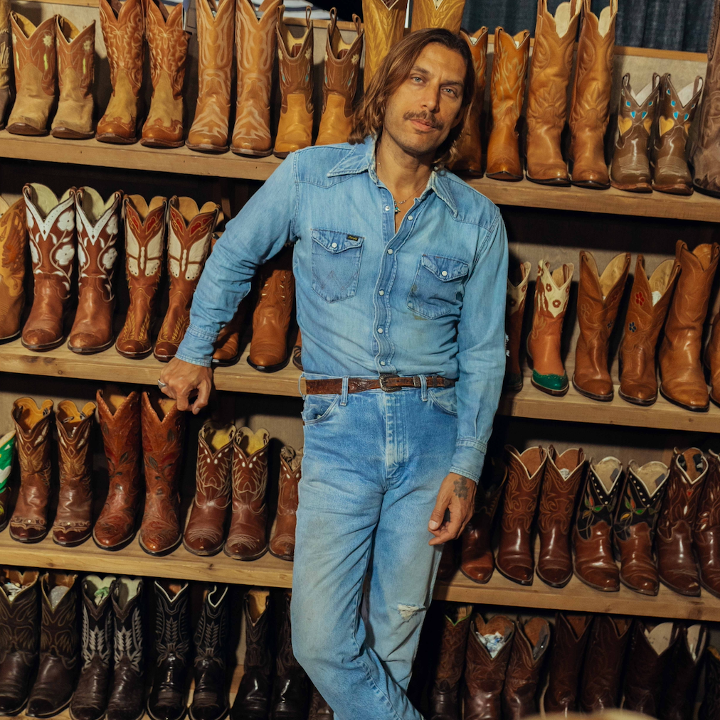 A man wearing a light blue denim shirt and matching jeans stands in front of shelves filled with cowboy boots in a rustic indoor setting.