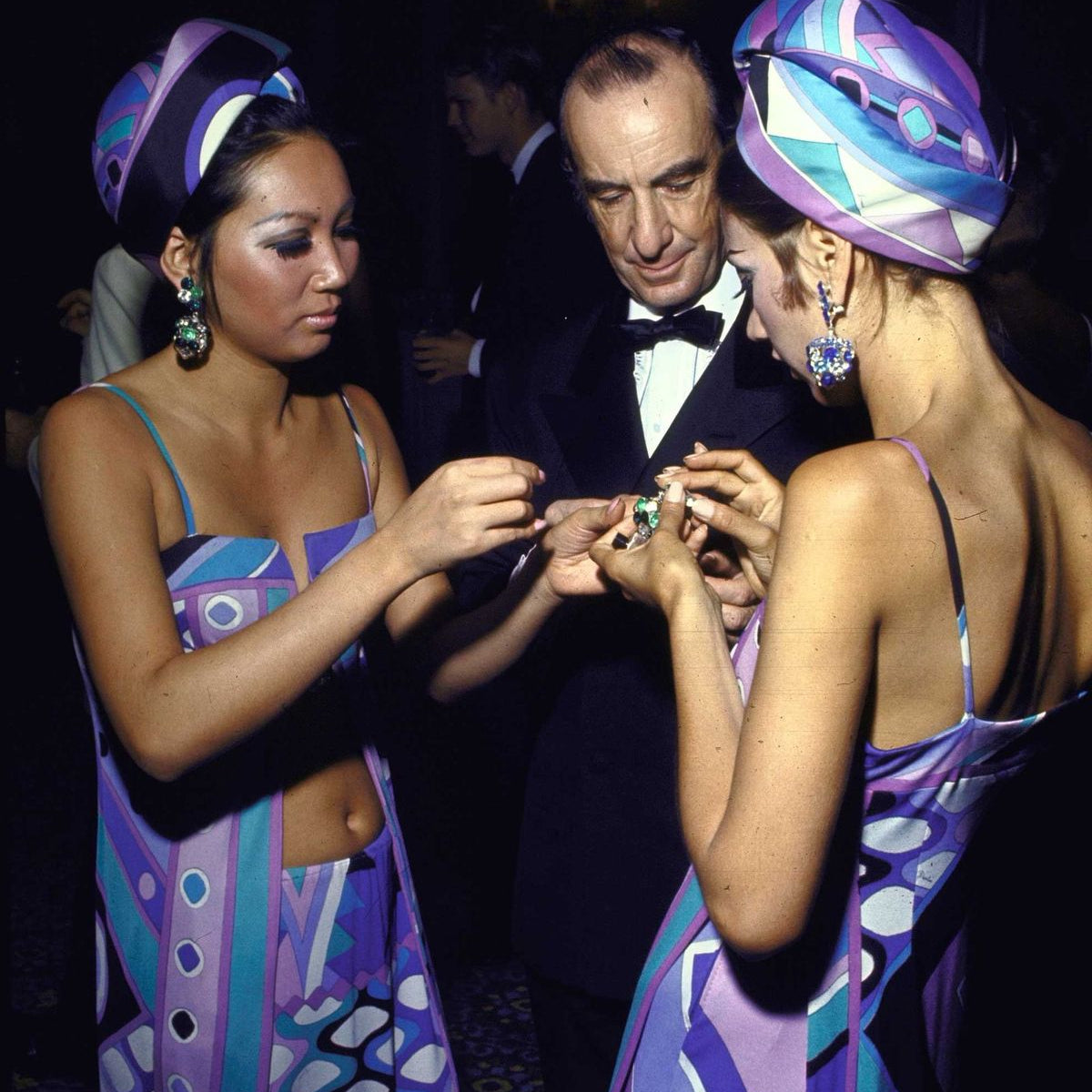 Two women in colorful dresses and headscarves interacting with a man in formal attire against a dark background.